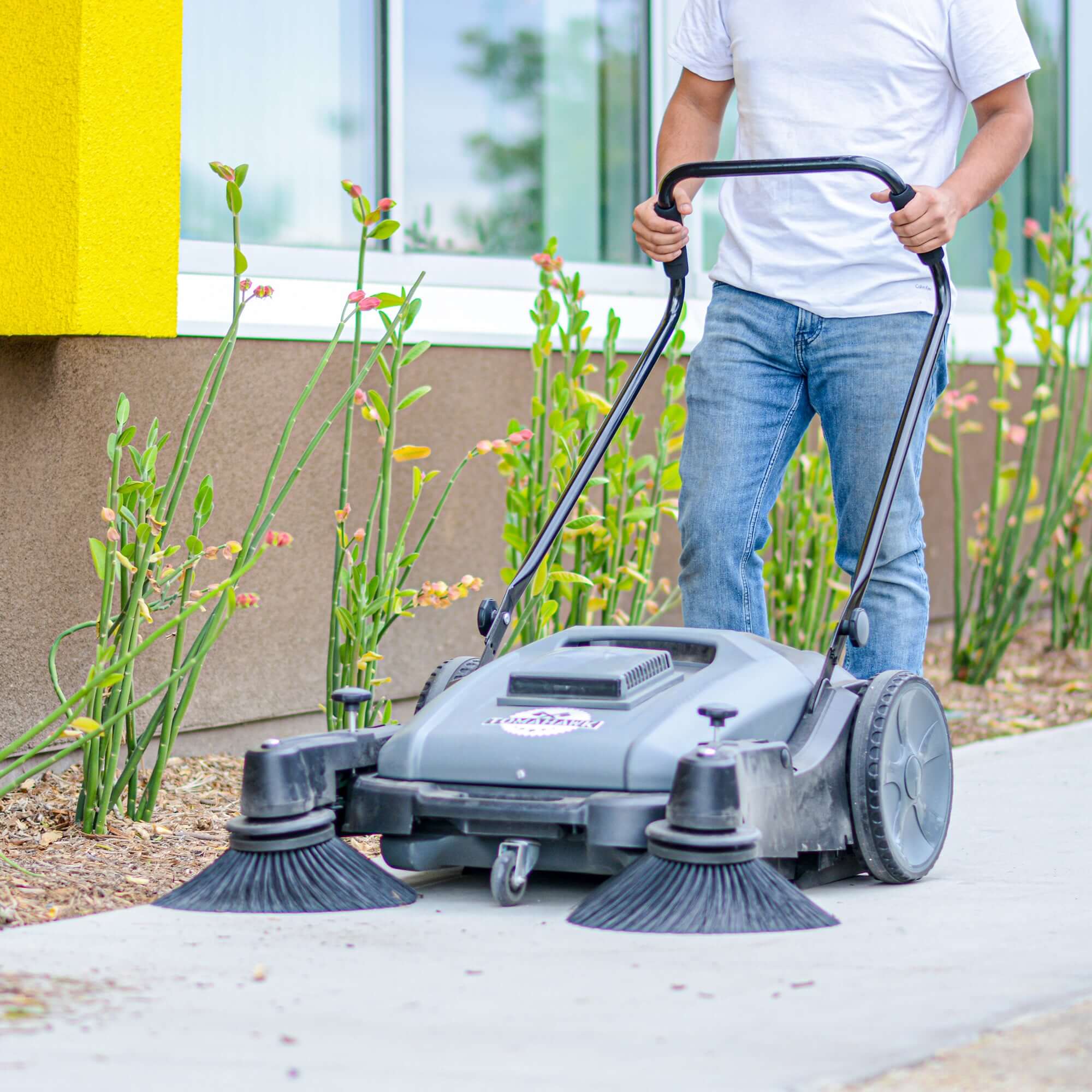 Man pushing a Tomahawk sweeper on a sidewalk outside.